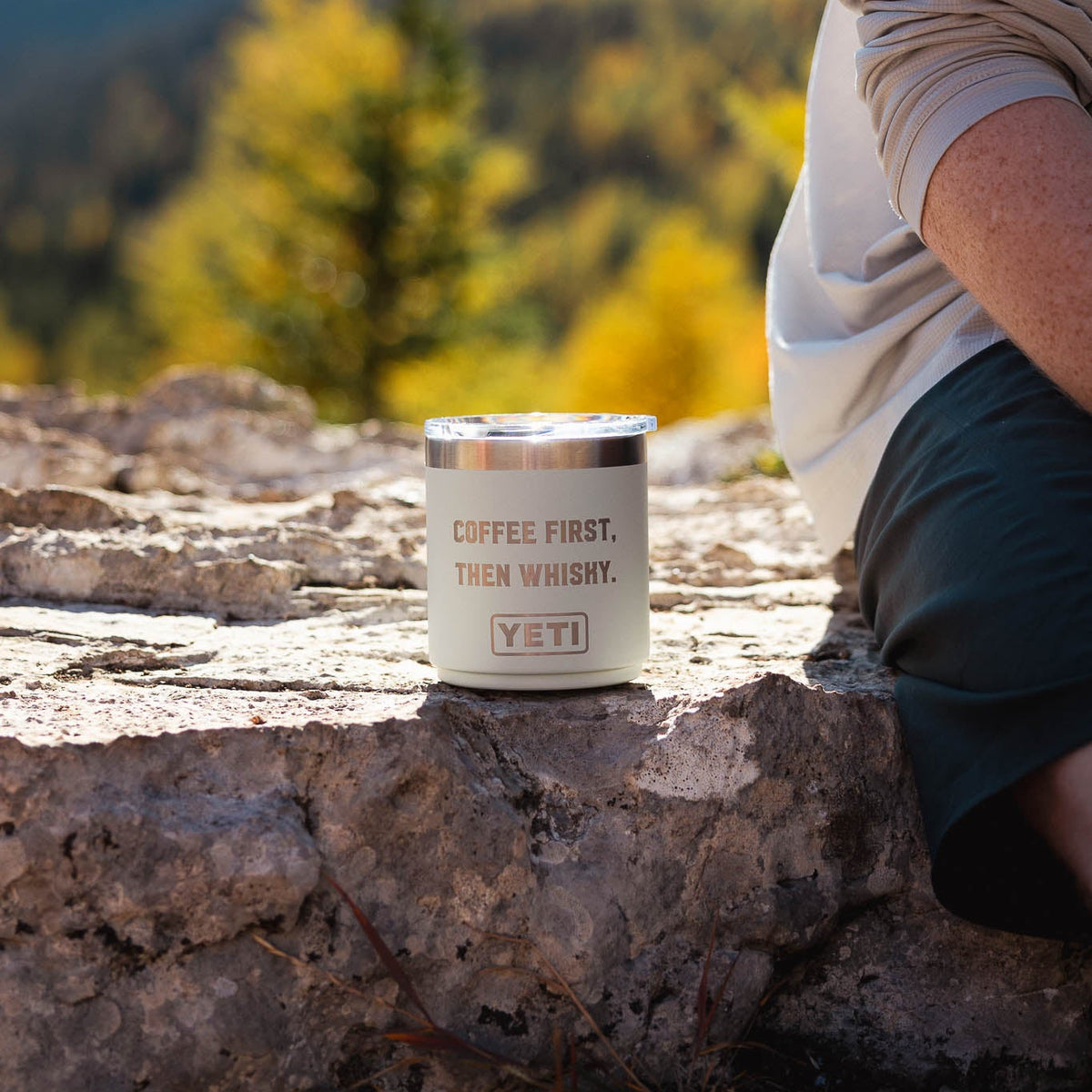 A light blue YETI insulated tumbler with a silver lid sits on a rocky outdoor ledge. The tumbler has the text 'COFFEE FIRST, THEN WHISKEY.